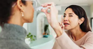 A woman using eye drops while looking in a mirror.