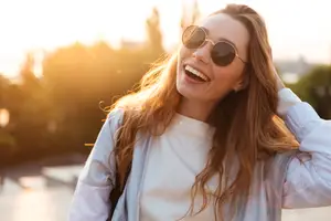 A woman with long hair smiling and wearing sunglasses, probably in a park during the daytime