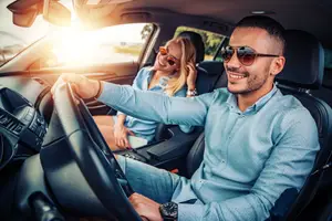 A man and a woman sitting inside a car, the man is driving while the woman is smiling and the man is wearing sunglasses and a bracelet.