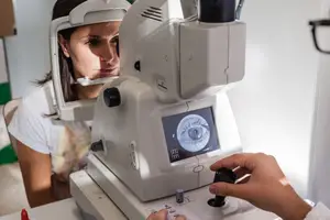 A woman is looking into a machine with a black button on it, and it seems to be an eye examination machine. A person is holding the button while looking at the monitor.