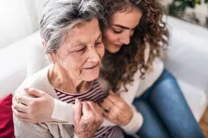 An elderly woman with gray hair is sitting on a white couch while holding the shoulder of a woman with curly hair who is sitting next to her and looking at her.