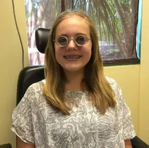 A young girl with blonde hair wearing a gray patterned blouse and large round glasses is smiling and posing for a photo in front of a window with a view of trees outside.