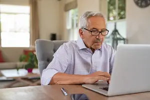 An older man is sitting on a chair in front of a desk, using a laptop, and wearing glasses.