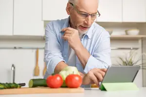 An older man with glasses and a blue shirt sits at a kitchen counter while looking at a tablet and chopping vegetables.