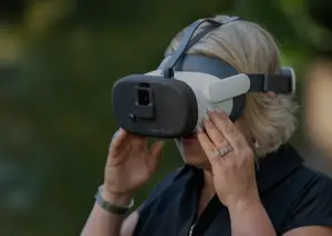 A woman wearing a black shirt and a black and white virtual reality headset is looking up with her right hand on her face.