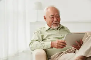 An older man sitting on a chair and looking at a tablet