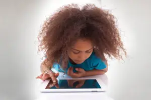 A child with curly hair is sitting and using a tablet, possibly in a room.