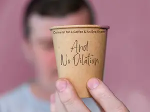 A person holding a coffee cup with a message that reads, 'Come In for a Coffee and an Eye Exam And No Vilation.'