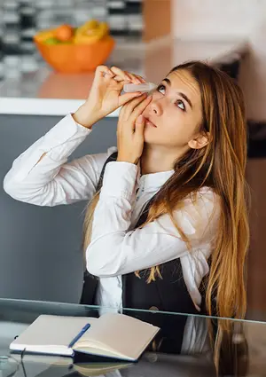 A young woman using eye drops in a kitchen.