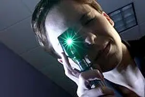 A man is looking through a green light ophthalmoscope with a smile on his face