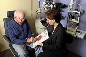 An elderly man wearing glasses examines an eye chart with an optometrist in an examination room.