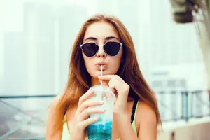 A young woman is wearing sunglasses and drinking a blue beverage from a plastic cup with a straw on a balcony.