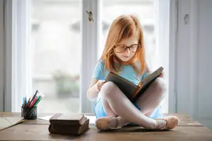 A girl reading a book while sitting on a wooden table