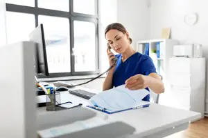 A woman with blue scrubs is answering a phone call while holding a piece of paper in her hand.