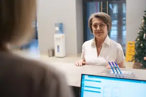 A woman with glasses and a white shirt is smiling and standing behind a desk with a computer monitor, a water dispenser, and a Christmas tree in the background.