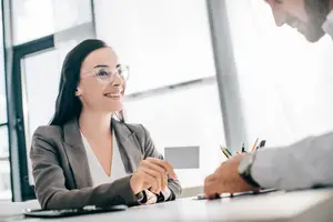 A woman with glasses is smiling and talking to a man in a suit in an office room