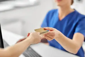 A woman in a blue scrub uniform stands behind a desk, handing over a card to a person in front of her in a room with a computer and keyboard.
