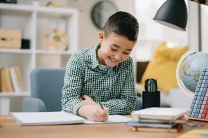 A smiling young boy is sitting at a desk with books and writing materials.