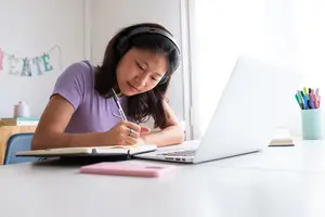 A young woman wearing headphones is sitting at a desk, writing in a notebook while wearing a purple shirt.
