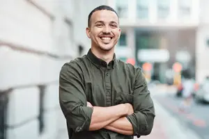 Man in green long sleeve shirt standing on street smiling with arms crossed