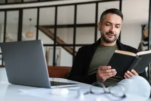 A man is reading a book while sitting on a chair in front of a desk with a laptop.