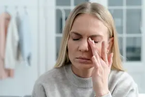 A woman with blonde hair is sitting on a couch and covering her face with her right hand. She is probably feeling sick or has a headache. She is looking at something in front of her, probably a TV or a laptop. Behind her, there is a white wall with a glass window. There are also clothes hanging on the hanger on the left side. In the distance, there is a blurry image of a pink cloth.