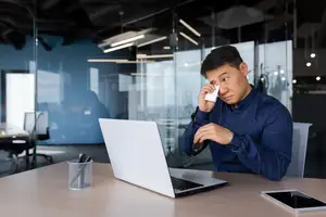 An adult male sitting at a desk in an office building looking at a laptop while holding a tissue to his face.