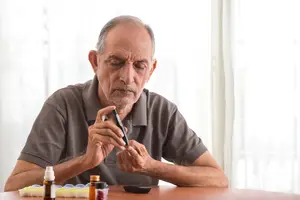An elderly man with gray hair, wearing a brown polo shirt, is sitting on a chair, checking his blood sugar with a glucometer while looking at it. He is sitting on a wooden table with a black object, a bottle, and a medicine organizer on top. Behind him is a white curtain.