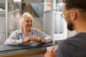 An elderly woman wearing glasses is smiling while sitting on a chair in front of a man wearing glasses. The man is holding a pen and paper, probably writing. He is probably a bank teller. Behind them is a glass wall with a blurry view of a building and a staircase.
