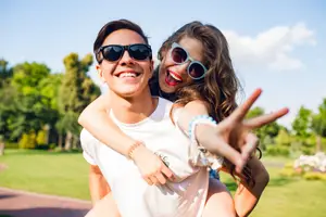 A man and a woman are smiling while posing for a photo in a park, the woman is making a peace sign with her hand