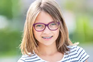 A young girl with braces and glasses is smiling and looking forward, possibly posing for a photo.