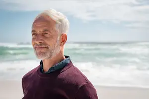A smiling man in a maroon sweater standing on a beach with the ocean in the background