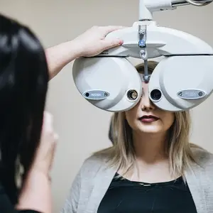 A woman with blonde hair is getting her eyes checked by an optometrist using a machine with the brand name NIDEX. The woman is wearing a gray cardigan and a black top. The optometrist is wearing a bracelet and a ring on her left hand.