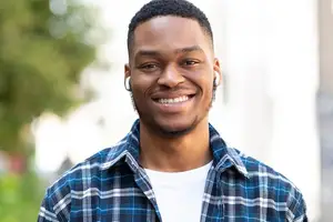 A young black man wearing a blue and white checkered shirt and white earphones with a smile on his face, standing on the street and posing for a photo