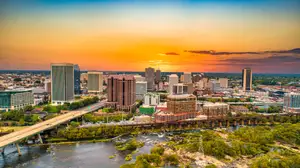 A city skyline with several high-rise buildings at sunset, including a bridge over a river and trees in the foreground.