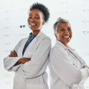 Two women in white lab coats standing next to each other in front of a wall with eyeglasses displayed on it.