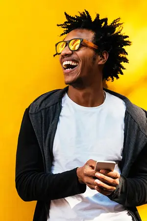 A smiling man wearing sunglasses and a white t-shirt is standing in front of a yellow wall and looking at his phone.