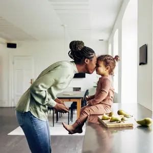 A mother and child are sitting on a kitchen counter, the mother is kissing the child on the cheek, the child is holding a pear and there is a wooden chopping board with pears on it.