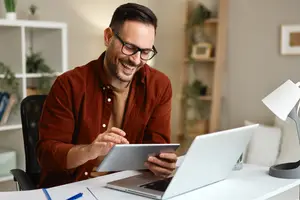 Man sitting at a desk using a tablet and laptop, smiling at the camera.