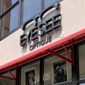 The front of an Eyesee Optique store with a red awning, featuring a black and white sign and a photo of a person wearing glasses.