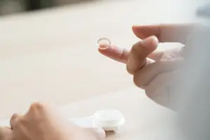 A person is holding a contact lens in their right hand and a contact lens container in their left hand on a wooden table.