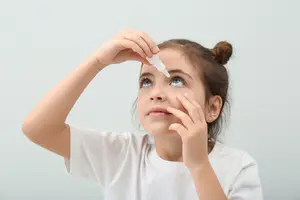 A young girl is administering eye drops while sitting on a couch, she has a serious expression on her face.