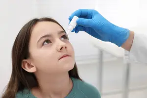 A girl with brown hair is looking at a bottle of eye drops being held by a person wearing blue gloves in what seems to be a clinic.