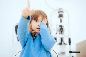 A girl is pointing at an eye exam machine while sitting in a chair in an ophthalmology clinic
