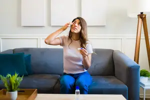 A woman sitting on a couch with a pillow and a potted plant on a table is putting in contact lenses and holding a bottle of contact lens solution.