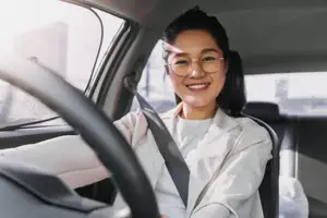 Photo of a smiling East Asian woman wearing glasses and driving a car with a seatbelt on
