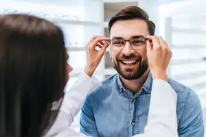 A man is getting his glasses adjusted by a woman in a white lab coat in a well-lit room.