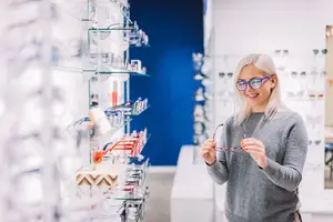 A smiling woman in a gray sweater holding a pair of glasses while looking at a display of glasses in a store.
