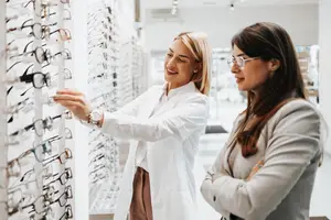 A woman in a white lab coat is showing another woman a display of glasses in a store.