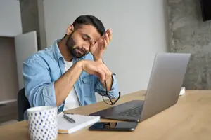 A man sitting at a desk with a laptop, holding his glasses, and looking tired